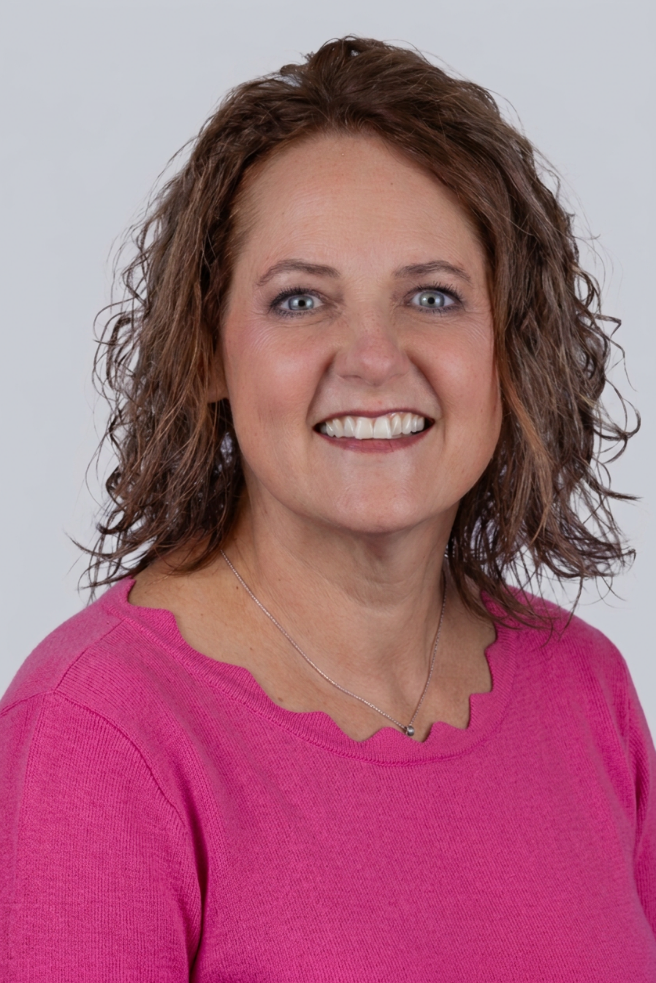 A smiling woman with curly hair wearing a bright pink top and a silver necklace, set against a plain white background.